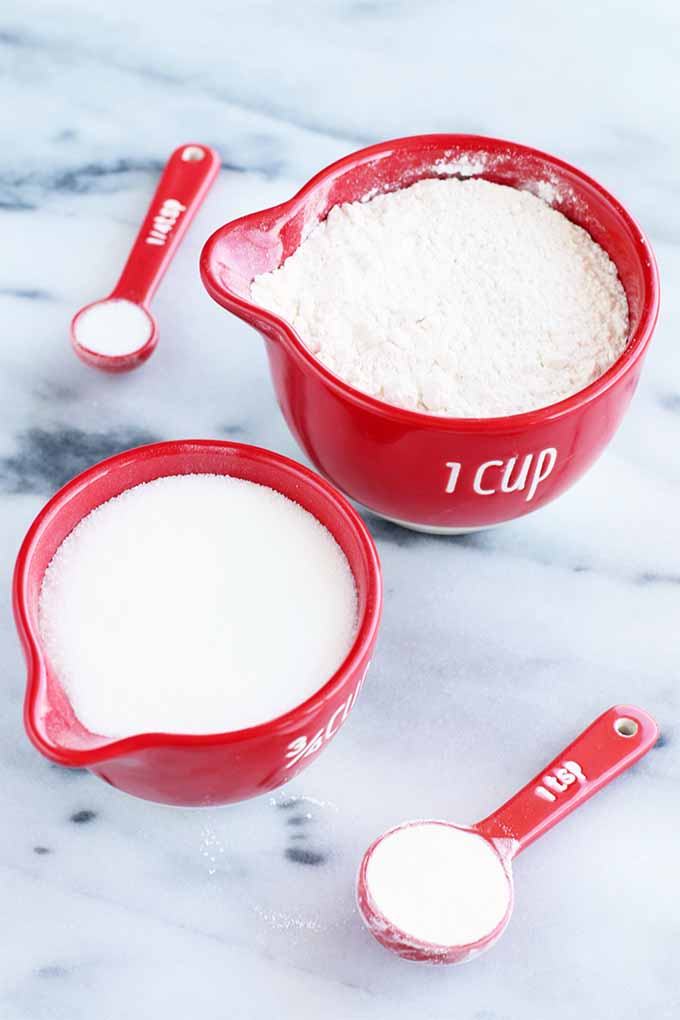 Red measuring cups with spouts filled with flour and sugar alongside red plastic measuring spoons of baking powder and salt, on a gray and white marble background.