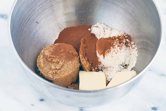 Packed brown sugar, cinnamon, flour, and butter in a stainless steel mixing bowl, on a marble background.