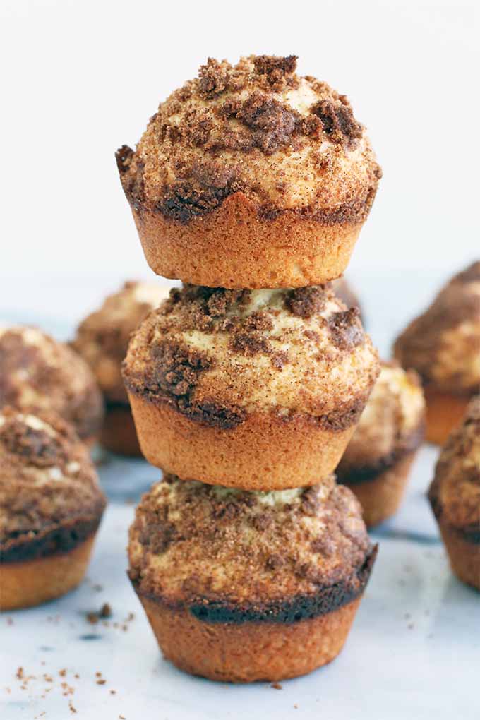 A vertical stack of three crumb cake muffins, with more in the background, on a marble surface with some cinnamon crumbs, in front of a white background.