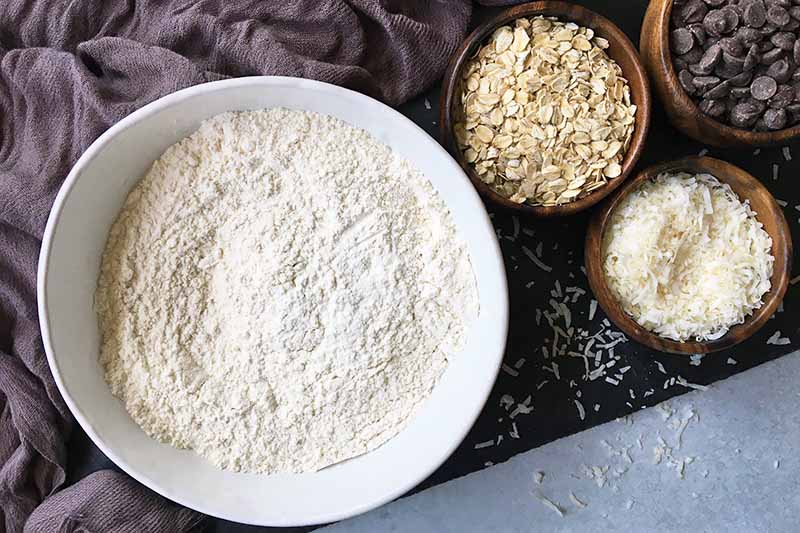 Horizontal image of a bowl with white dry ingredients next to wooden bowls with oats and shredded coconut.