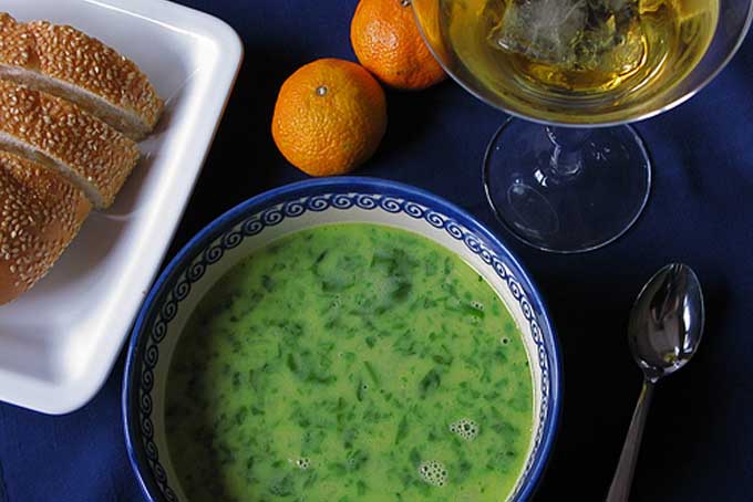 Top down view of a green bowl of spinach soup along with a loaf of bread, two oranges, and glass of wine | Foodal