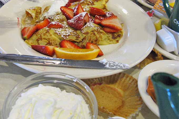 A plate of pancakes and a bowl of whipped cream on a table with packets of butter, a muffin wrapper, and other breakfast items, at Omega Restaurant in Illinois.