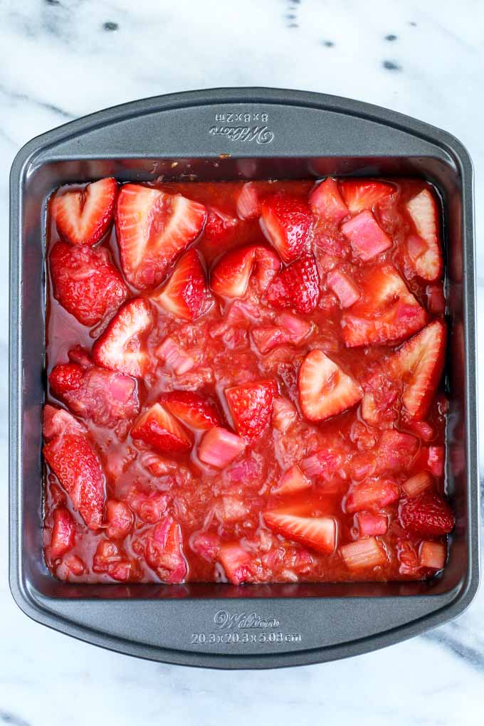 Top-down view of a square metal baking pan filled with cooked rhubarb, red syrup, and halved fresh strawberries, on a white and gray marble surface.