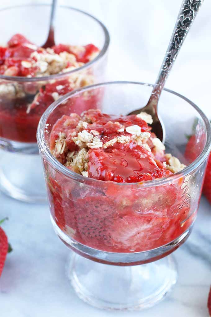 Two parfait glasses of rhubarb strawberry crumble dessert with silver spoons stuck into the cups and scattered whole strawberries, on a gray marble surface.