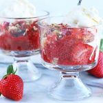 Rhubarb strawberry crumble in two parfait glasses, topped with vanilla ice cream with silver spoon, on a gray piece of marble with scattered whole strawberries and a white cloth.