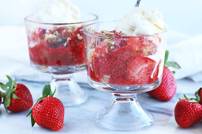 Rhubarb strawberry crumble in two parfait glasses, topped with vanilla ice cream with silver spoon, on a gray piece of marble with scattered whole strawberries and a white cloth.