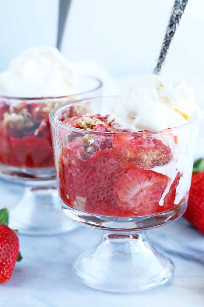 Two parfait glasses of strawberry rhubarb crumble topped with vanilla ice cream with spoons stuck into the glasses, on a marble surface with scattered whole strawberries.