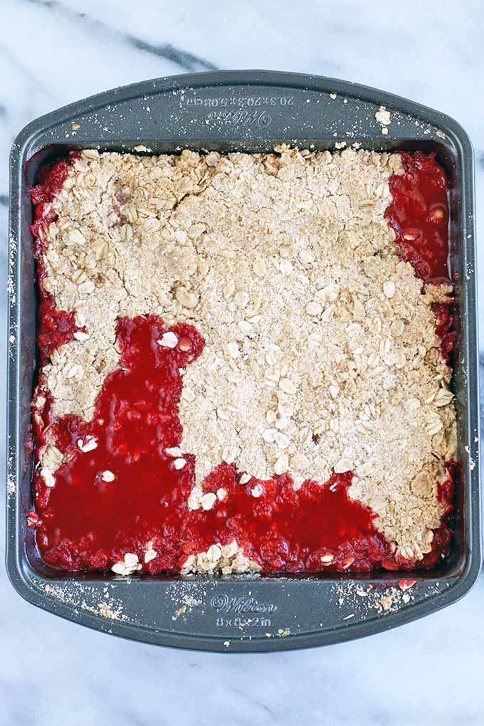 Top-down view of a square metal baking pan of red fruit crumble, on a gray and white marble slab.