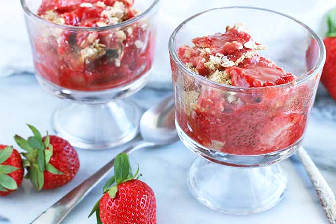 Two parfait glasses of cooked rhubarb and strawberries with a brown sugar and oat crumble topping, with a spoon and three whole strawberries on a marble slab.