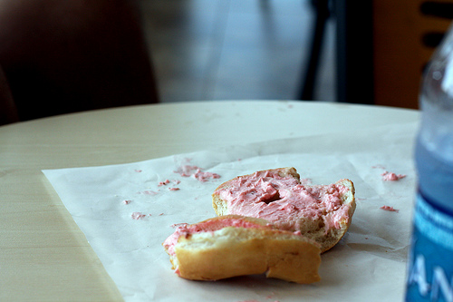 A bagel with pink frosting on a tissue on top of a table. 
