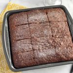 Horizontal image of a square pan with a baked chocolate cake lightly dusted in powdered sugar on a yellow towel next to a metal spatula.