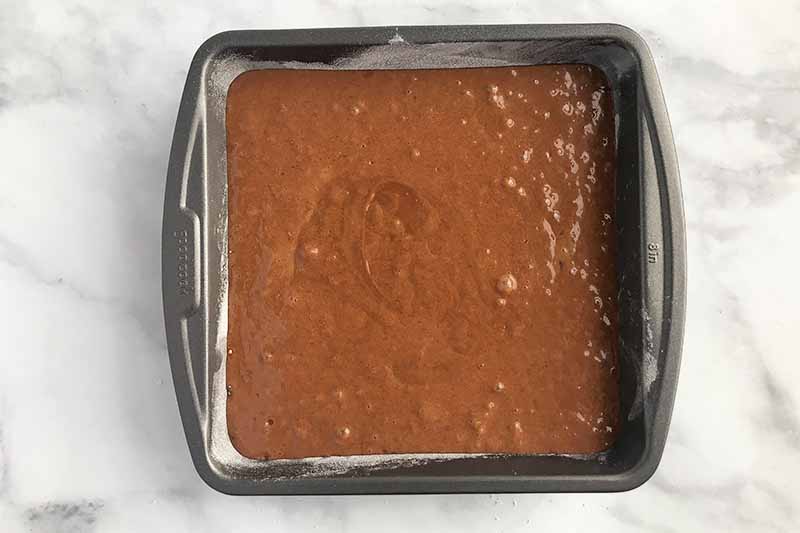 Horizontal image of a dark brown batter in a square pan on a white and gray surface.