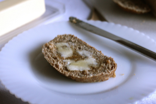 A close up image of white plate with a slice of whole grain bread with butter spread on it and a bread knife beside it.