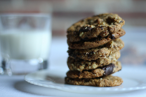 A stack of delicious looking dark cookies on a white plate and glass of milk at the back.
