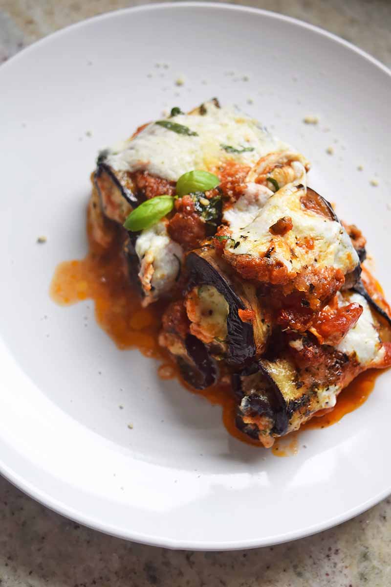 Vertical image of a white plate with a large serving of eggplant rolls with cheese, basil, and marinara.