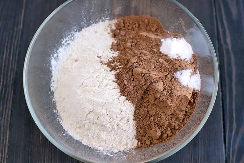 Overhead shot of flour, cocoa powder, salt, and baking powder in a large stainless steel mixing bowl, on a dark brown wood surface.