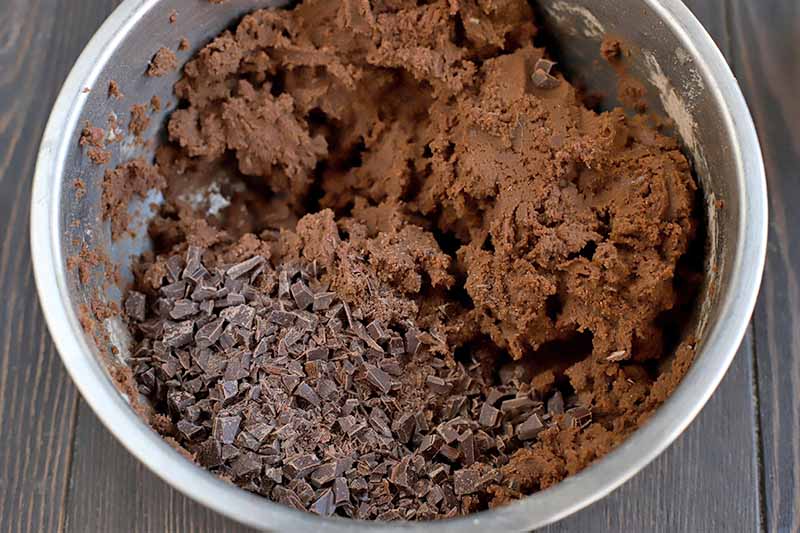 Cookie dough topped with chopped chocolate in a large stainless steel mixing bowl, on a dark brown wood background.