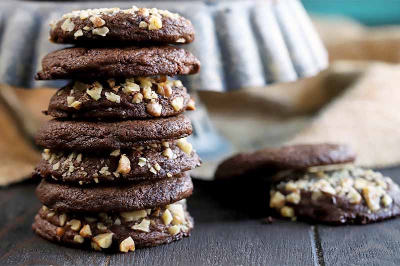 A stack of walnut-coated chocolate cookies on a dark brown wood surface, to the left of a few more on soft focus to the right, with a ceramic cake stand and a piece of burlap in the background.