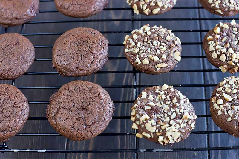 Plain and walnut-coated chocolate cookies cooling on a black metal rack, on a dark brown wood surface.
