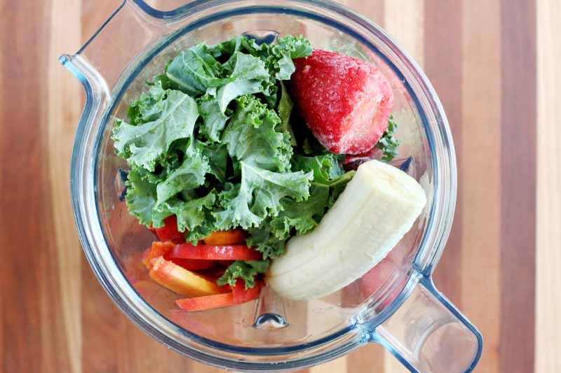 A plastic blender pitcher if filled with torn kale, sliced peaches, frozen strawberries, and a banana, on a striped brown wood surface.