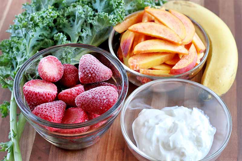 Three small glass bowls of frozen whole strawberries, sliced peaches, and Greek yogurt, with a few leaves of green kale and a yellow banana.