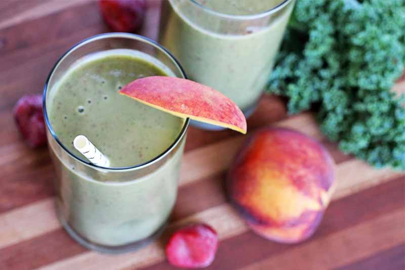 Two glasses of green smoothie with a white paper straw and a slice of peach on the rim, with a whole stone fruit, frozen berries, and a large leaf of green kale with curly edges, on a brown striped wood table.