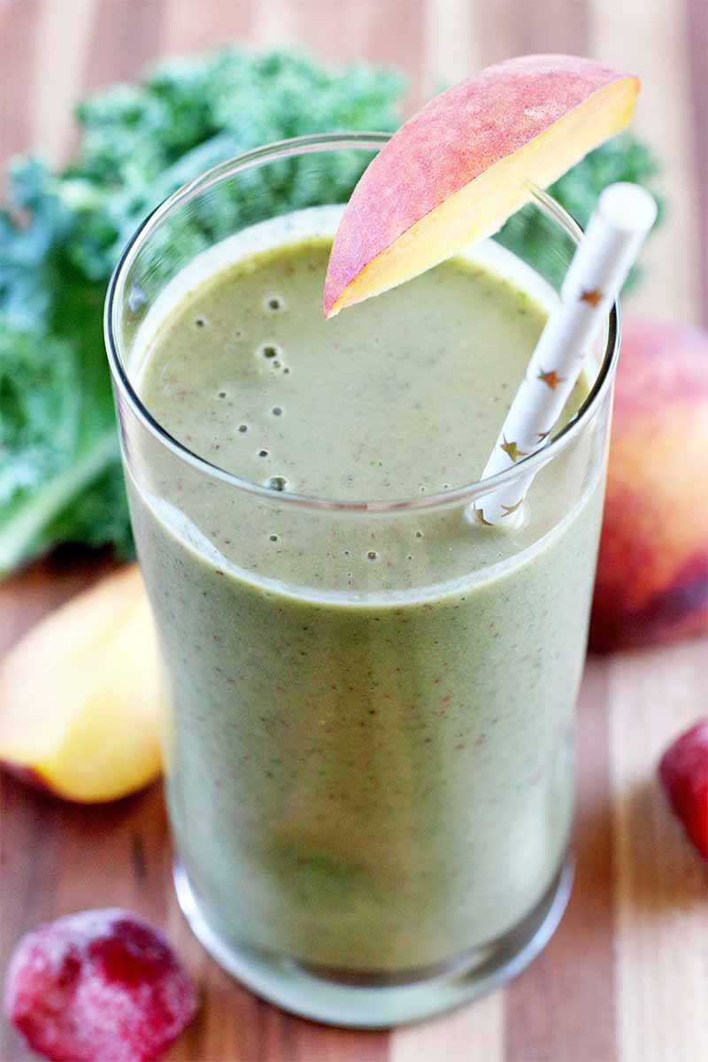 A tall glass if filled with a pale green smoothie, with a slice of peach with the skin on for garnish, and a gold and white paper straw, on a striped wood surface with more stone fruit, frozen strawberries, and curly-leaf kale.