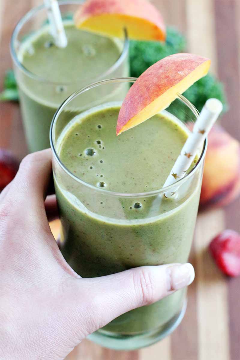 A woman's hand holds a glass of green smoothie with a peach slice for garnish and a white and gold paper straw, with another glass in the background, and a whole stone fruit, frozen strawberry, and leaf of green kale in the background in soft focus, on a striped wood surface.
