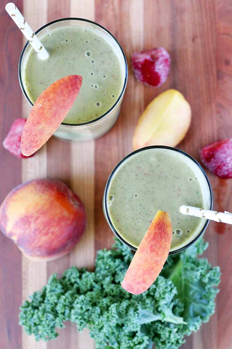 Top-down shot of two glasses filled with green smoothies with peach slices for garnish and white paper straws, surrounded by more stone fruit, frozen strawberries, and kale, on a brown wood surface.