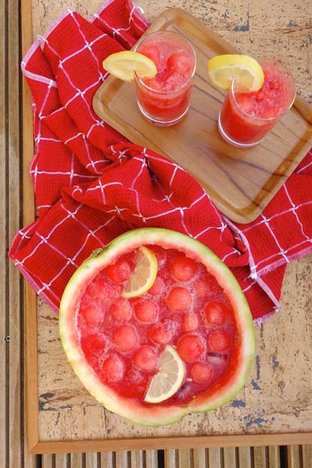 Overhead image of watermelon punch served in a hollowed out fruit, garnished with slices of lemon, with balls of melon and two full glasses on a small brown tray, on a red and white kitchen towel on top of a brown surface.
