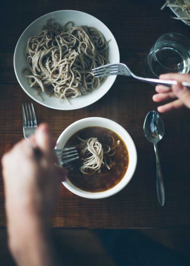 A bowl of soba noodles and a bowl of soup base with a pair of human hands adding noodles to the soup.