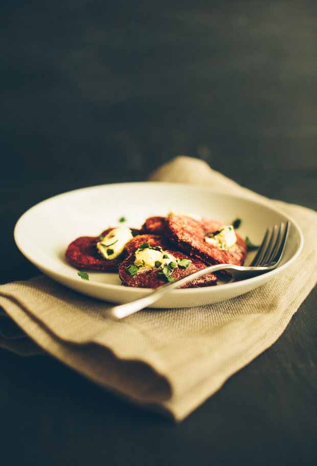 Vertical image of a shallow white ceramic bowl of heart-shaped beetroot patties topped with butter and chopped leafy greens, with a fork to the right, on a folded beige cloth napkin, on a black background.
