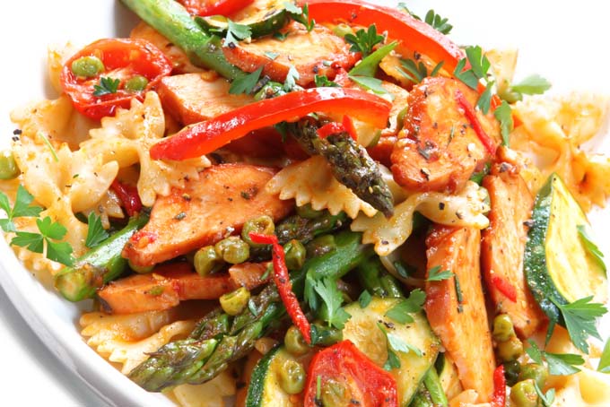 Horizontal image of a farfalle, mixed vegetable, and chicken dish on a white plate garnished with fresh herbs.