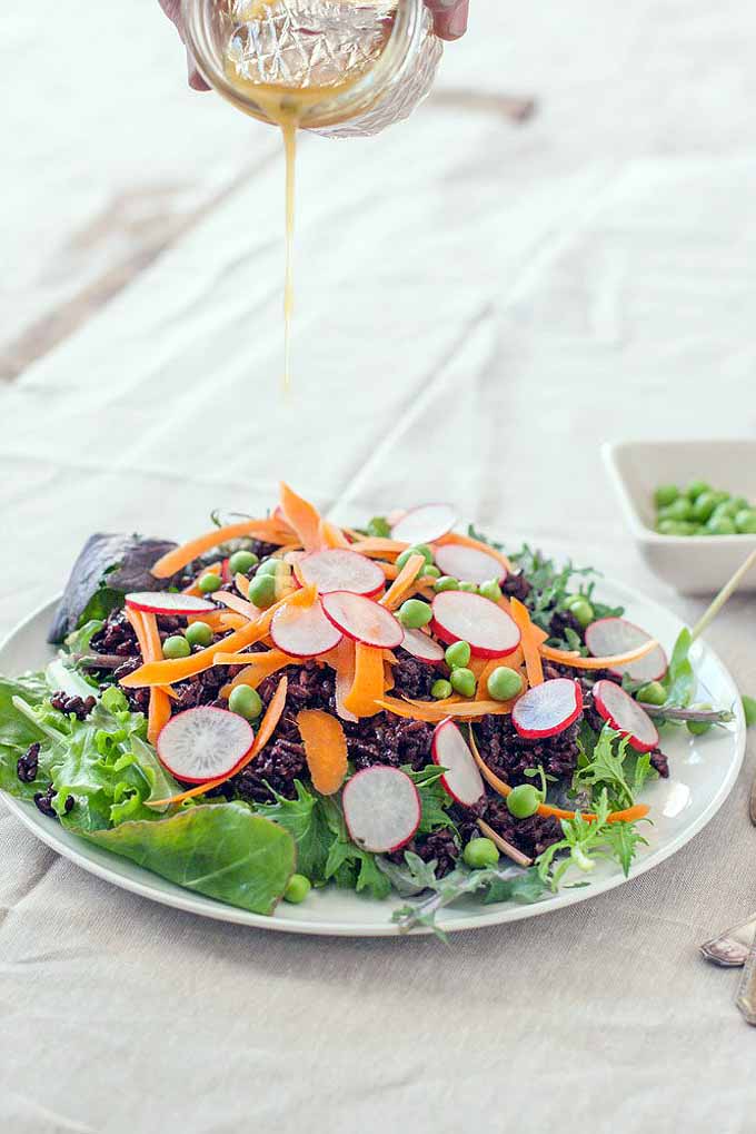 An oblique view of a black rice and pea salad with fresh veggies such as green lettuce, radishes, and shredded carrots on a white plate sitting on a white wooden table.
