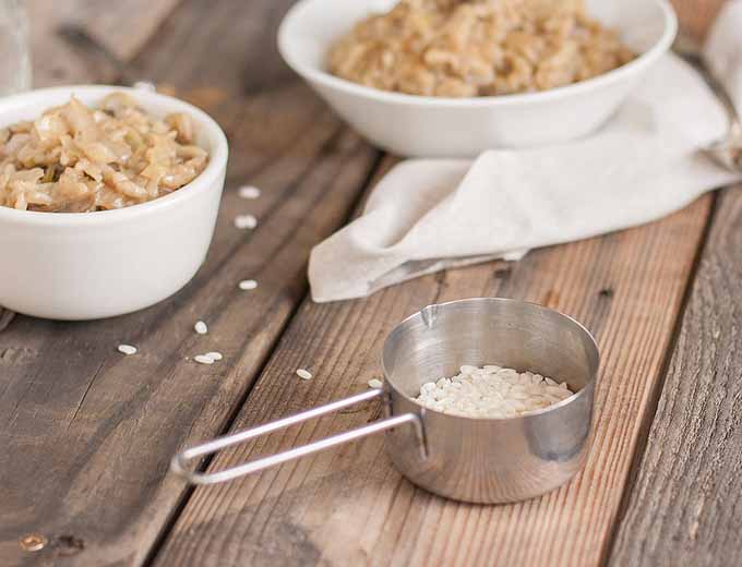 A measuring cup full of uncooked rice is in the foreground and two bowls of cabbage mushroom risotto is in the background. Everything is sitting on a unfinished wooden plank surface.