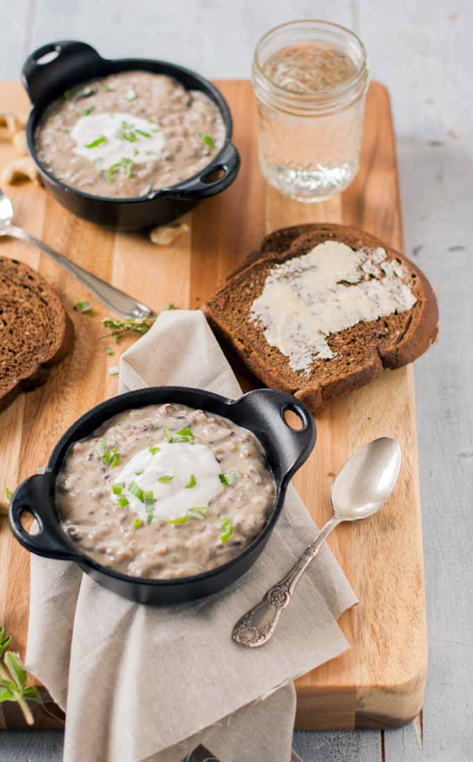 Creamy vegan wild rice and mushroom soup in two dark brown clay bowls with slices of dark bread sitting on a maple wood serving tray.