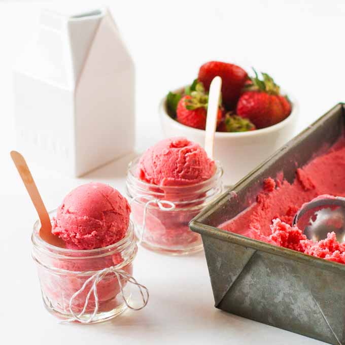 Oblique angle of two clear serving cups and a metal tray full of Strawberry and Goat Cheese Sherbet. A white porcelain cylinder shaped dish full of fresh berries is in the background.