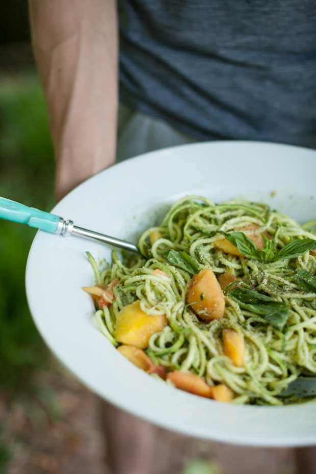 A bowl of zucchini spaghetti with peaches and pumpkin seed pesto being held in human hands.