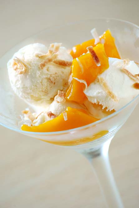 Vertical image of a martini glass filled with scoops of mango ice cream garnished with toasted coconut shreds on a light tan background.