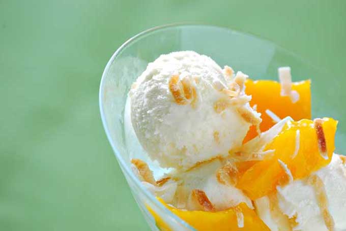 Horizontal image of a glass bowl with mango ice cream garnished with coconut shreds in front of a green background.