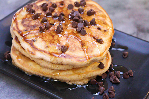 Horizontal image of a stack of cinnamon pancakes topped with maple syrup and a scattering of chocolate chips on a black plate.