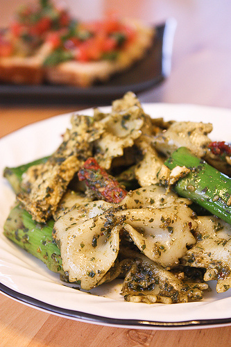 Vertical image of a white plate with farfalle pasta, asparagus, sun-dried tomatoes, and chicken pieces coated in pesto on a wooden table.