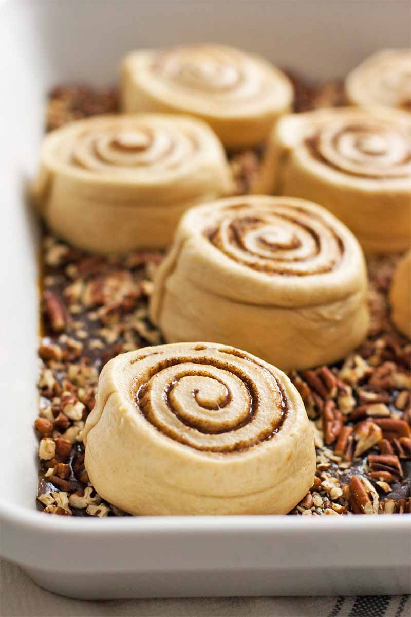 Round roll dough with a brown cinnamon spiral, in a white ceramic dish of pecans and brown sugar sauce.