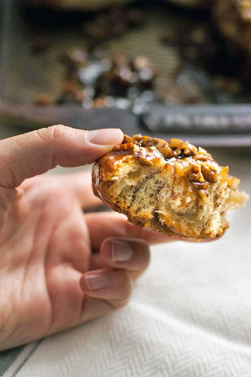 A hand holds half of a sticky bun up to the camera, with a kitchen in soft focus in the background.