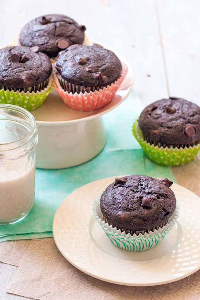 Vertical image of three vegan double chocolate muffins in pink and green paper wrappers on a white pedestal-style cake stand, with another muffin on a small white plate and one sitting directly on the white wooden table, with a folded pale aqua cloth and a mason jar of almond milk.