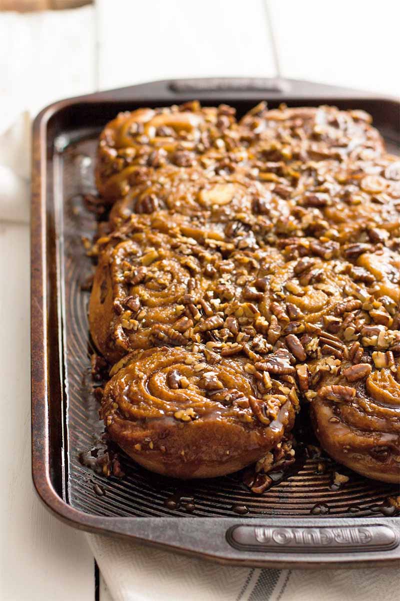 Pecan sticky buns fresh out of the oven, on a brown metal baking sheet, on a white tile background with a white cloth napkin that has a pale blue stripe.