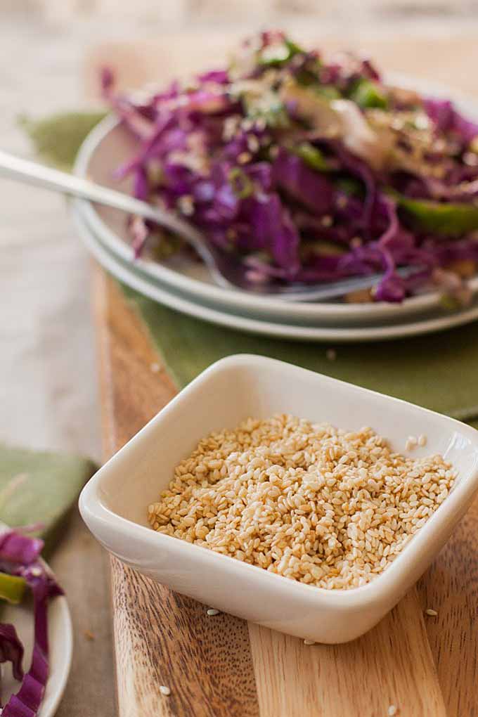 Closeup of a rectangular bowl of untoasted sesame seeds with a completed slaw in the background.