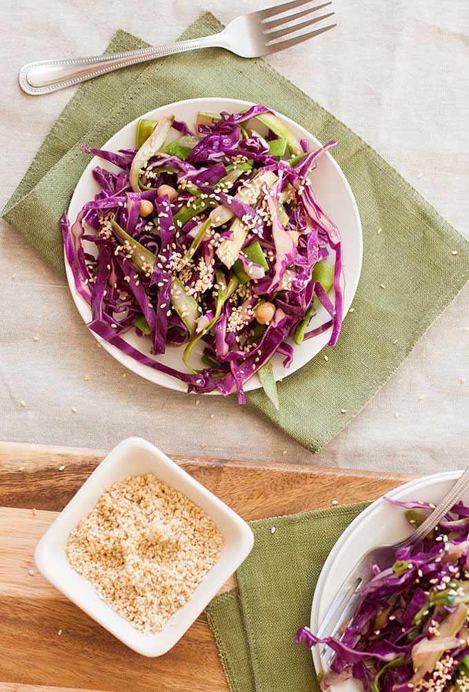 Top-down view of a plate full of purple cabbage and asparagus slaw. It is sitting on a green linen cloth place mat. A rectangular bowl of sesame seeds is in the lower left corner and a portion of a second bowl of slaw is visible in the lower right corner.