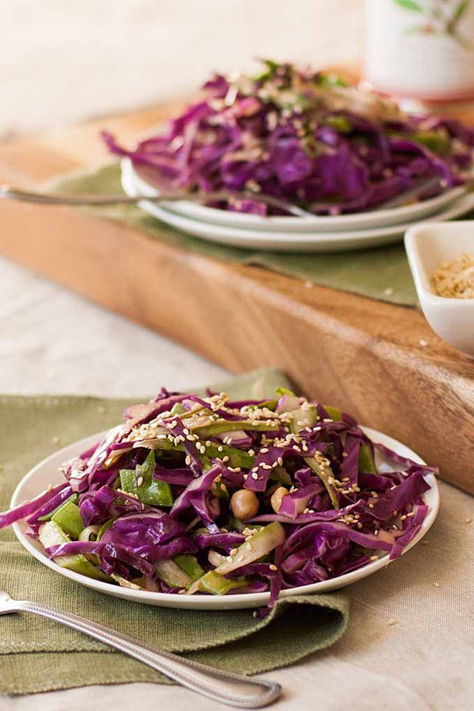 An oblique profile of two plates full of purple cabbage slaw. One is in the foreground on a green linen table setting and the other is in shallow focus in the background on a thick cutting board.