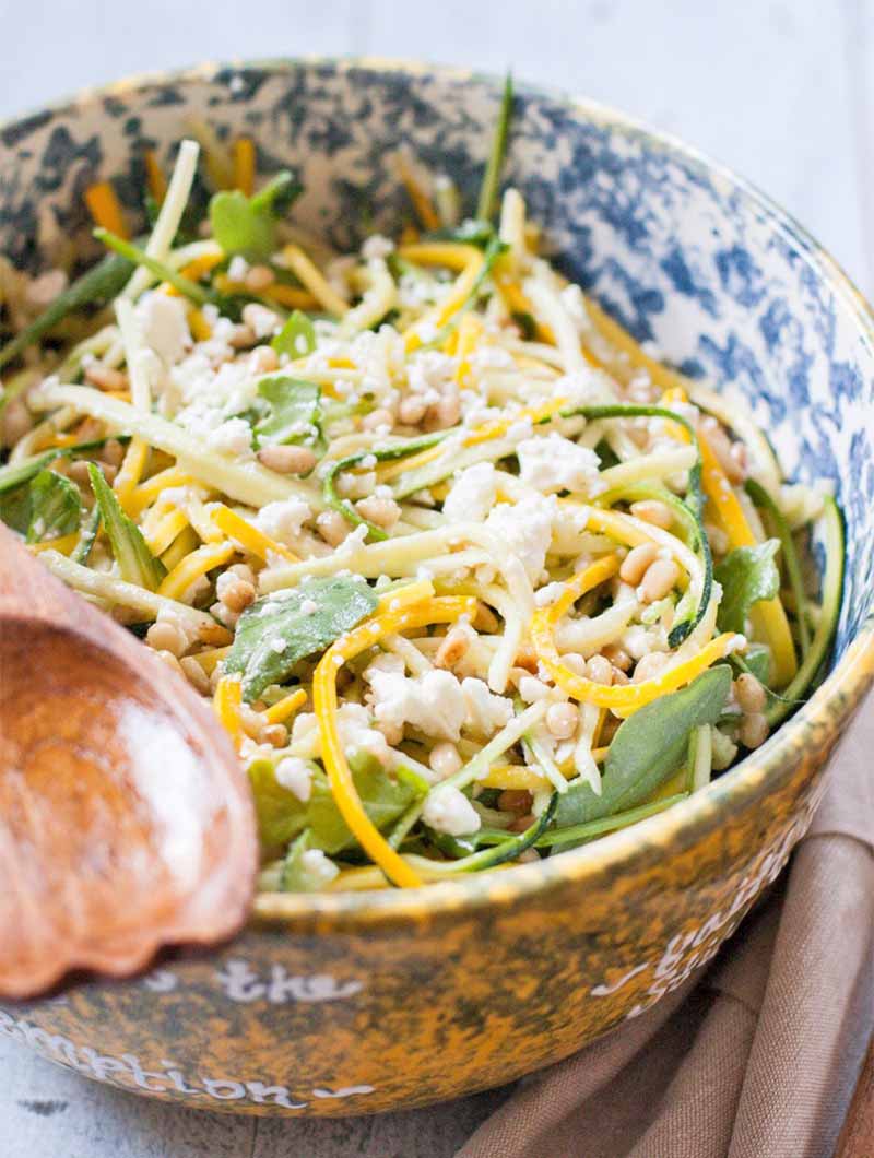 A yellow, blue, and white speckled ceramic bowl of summer squash salad with feta cheese, pine nuts, and arugula, with a wooden serving utensil and a folded beige cloth.